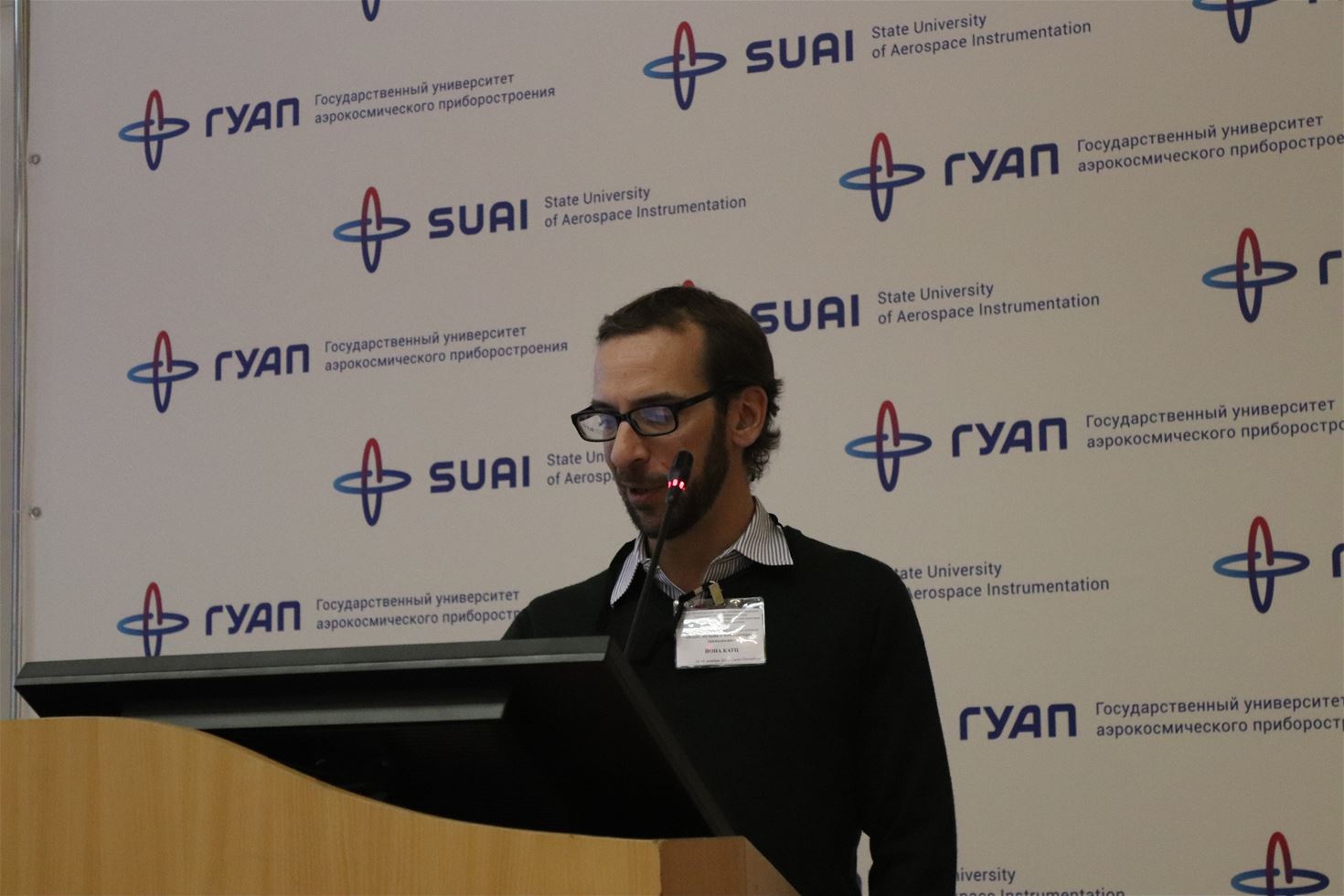 Jonah Katz wearing a name tag and a black sweater and striped collared shirt speaking at a podium.