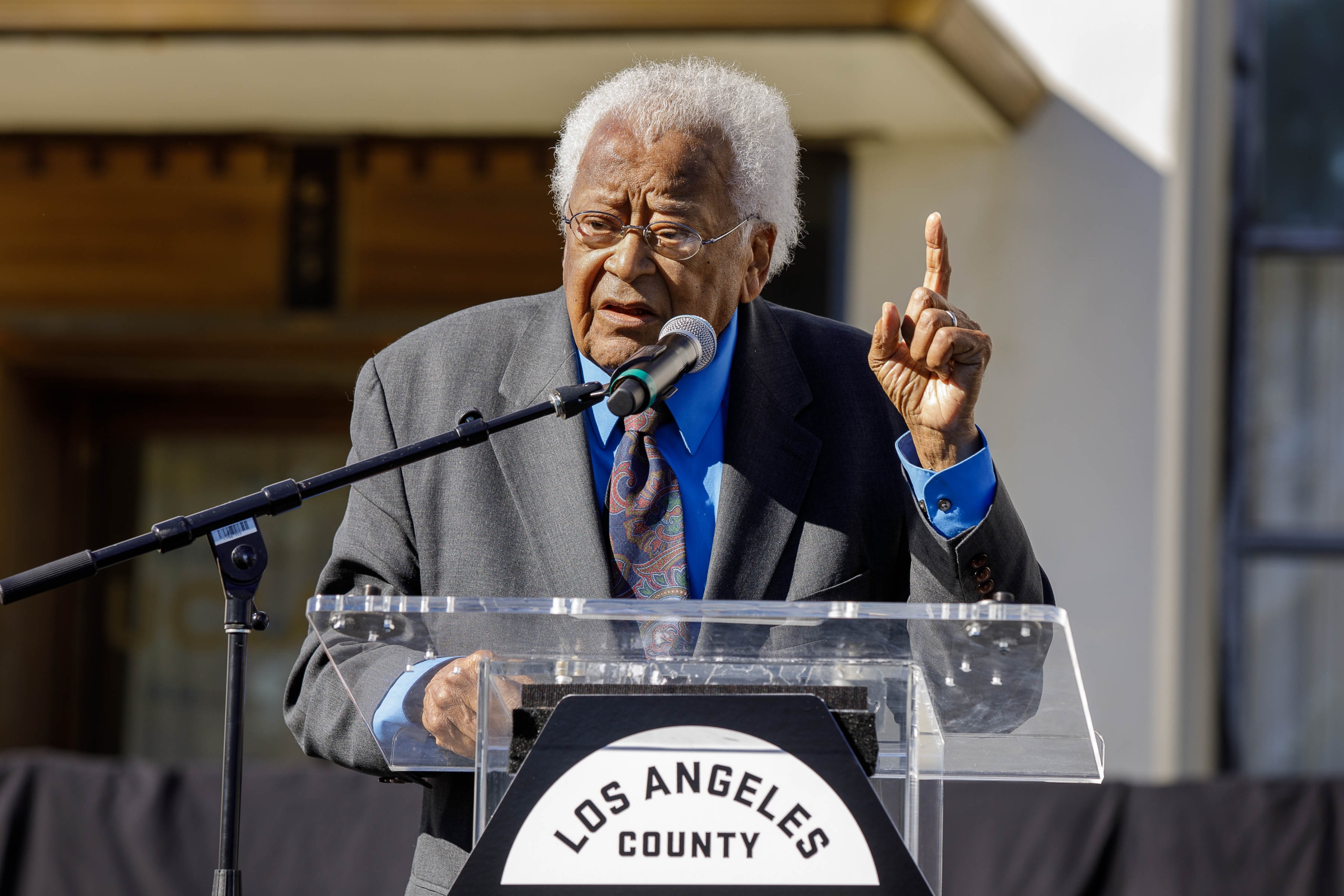 The Rev. James Lawson Jr. at building dedication speaking at a podium with a microphone in front of him and his left index finger raised.