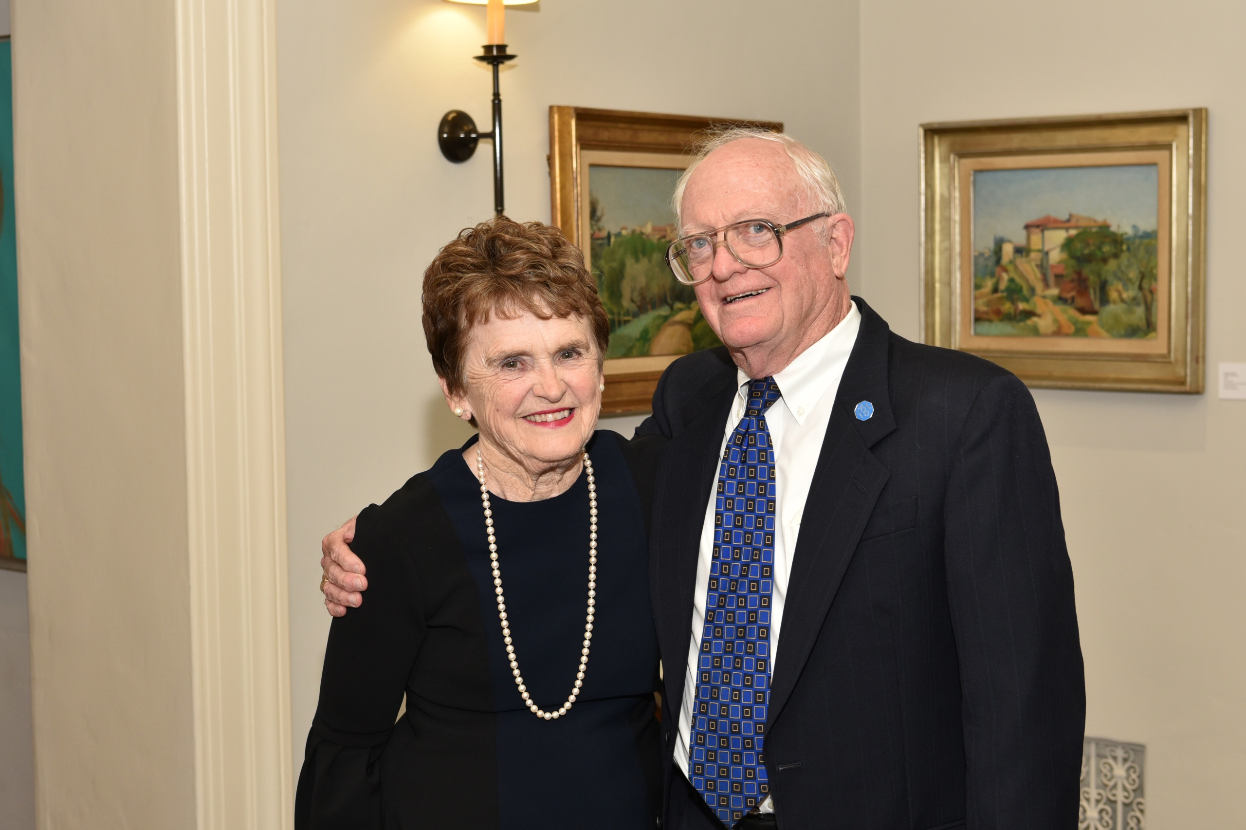 Sherie Morrison in a black dress and pearls stands next to Don Morrison in a black suite with a white shirt and blue and black tie; they stand in front of two paintings and a light.