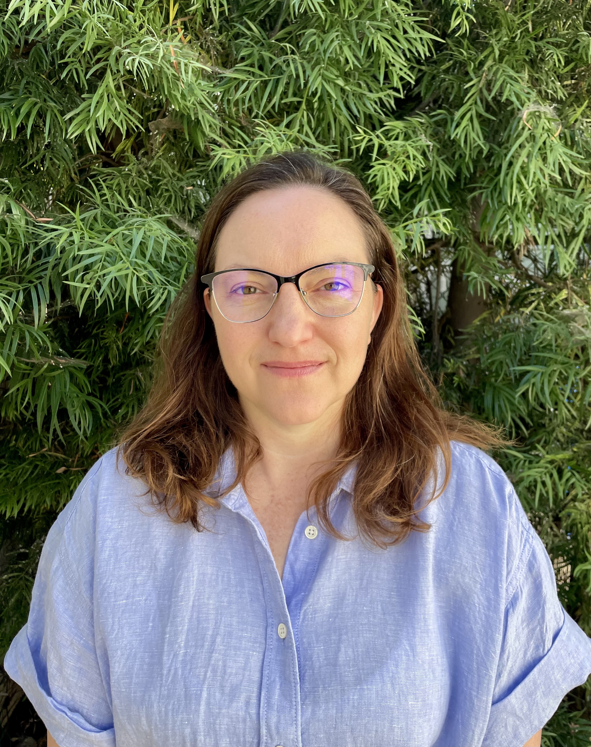 Nina Duthie in a blue shirt and glasses in front of some greenery.