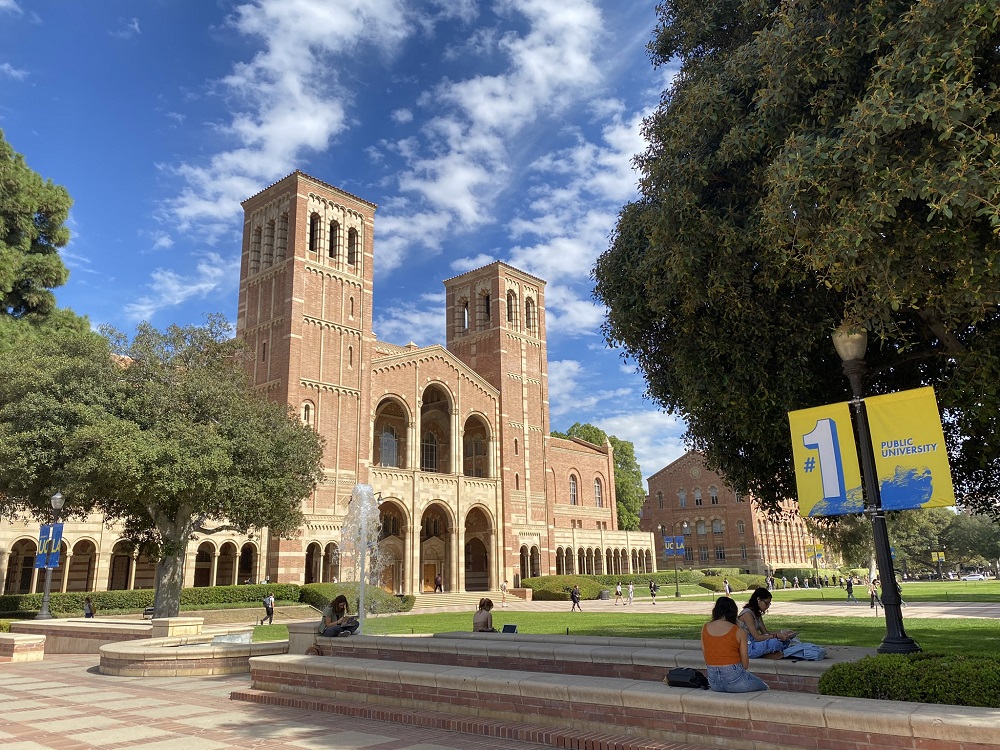 An image of UCLA's Royce Hall, Shapiro Fountain and Dickson Plaza with students sitting in the foreground, under a blue sky with scattered white clouds