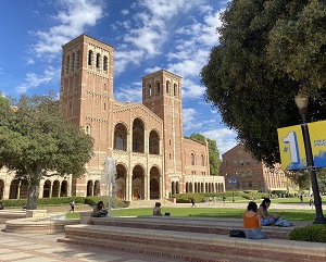 An image of UCLA's Royce Hall, Shapiro Fountain and Dickson Plaza with students sitting in the foreground, under a blue sky with scattered white clouds