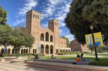 An image of UCLA's Royce Hall, Shapiro Fountain and Dickson Plaza with students sitting in the foreground, under a blue sky with scattered white clouds