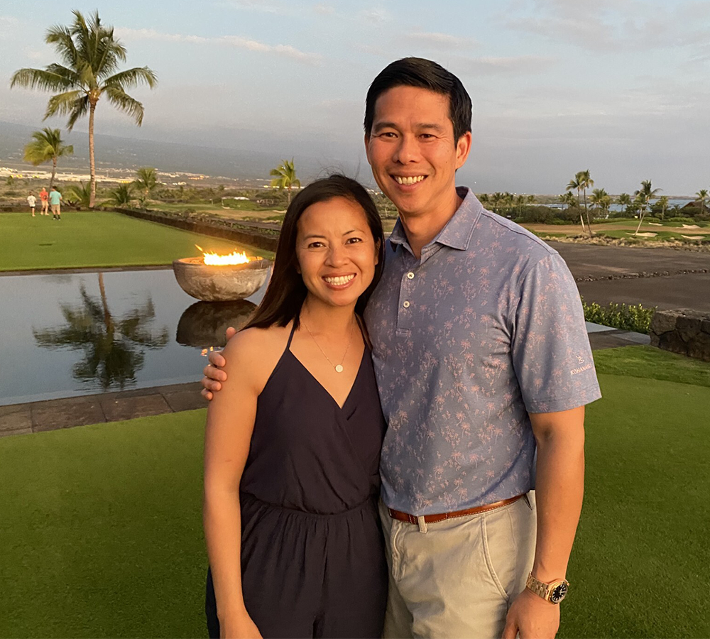 Pauline Le and Kiet Lam posing for a photograph on a green lawn with palm trees and a pond in the background. 