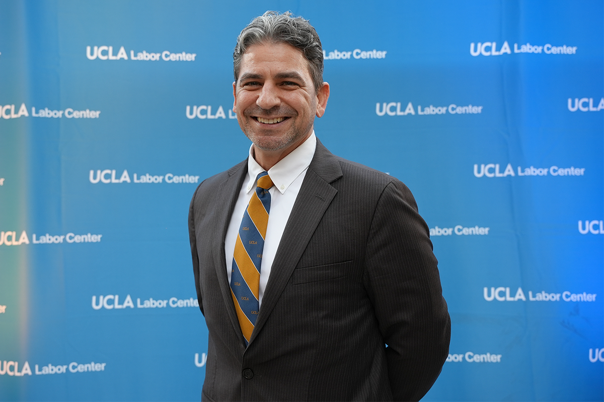 Nathan Seidman in a black suit with a blue and yellow tie in front of a blue background that reads, 