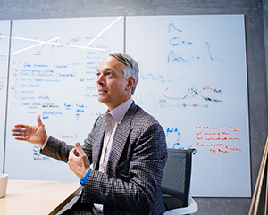 Alexander Hoffmann, in checkered suit, gestures as he sits at table in front of a whiteboard