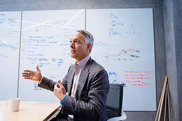 Alexander Hoffmann, in checkered suit, gestures as he sits at table in front of a whiteboard