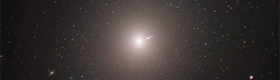 Surrounded by stars and galaxies, the galaxy M87, with a linear “jet” emitting from the center, is seen from a distance as captured by the Event Horizon Telescope.