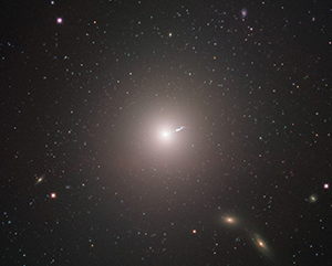 Surrounded by stars and galaxies, the galaxy M87, with a linear “jet” emitting from the center, is seen from a distance as captured by the Event Horizon Telescope.