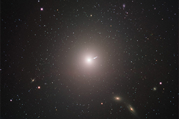 Surrounded by stars and galaxies, the galaxy M87, with a linear “jet” emitting from the center, is seen from a distance as captured by the Event Horizon Telescope.