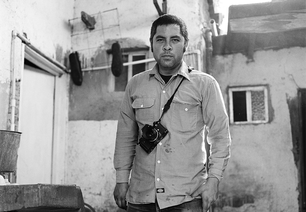 Black and white photo of Jason De León standing with a camera on a strap around his shoulder.