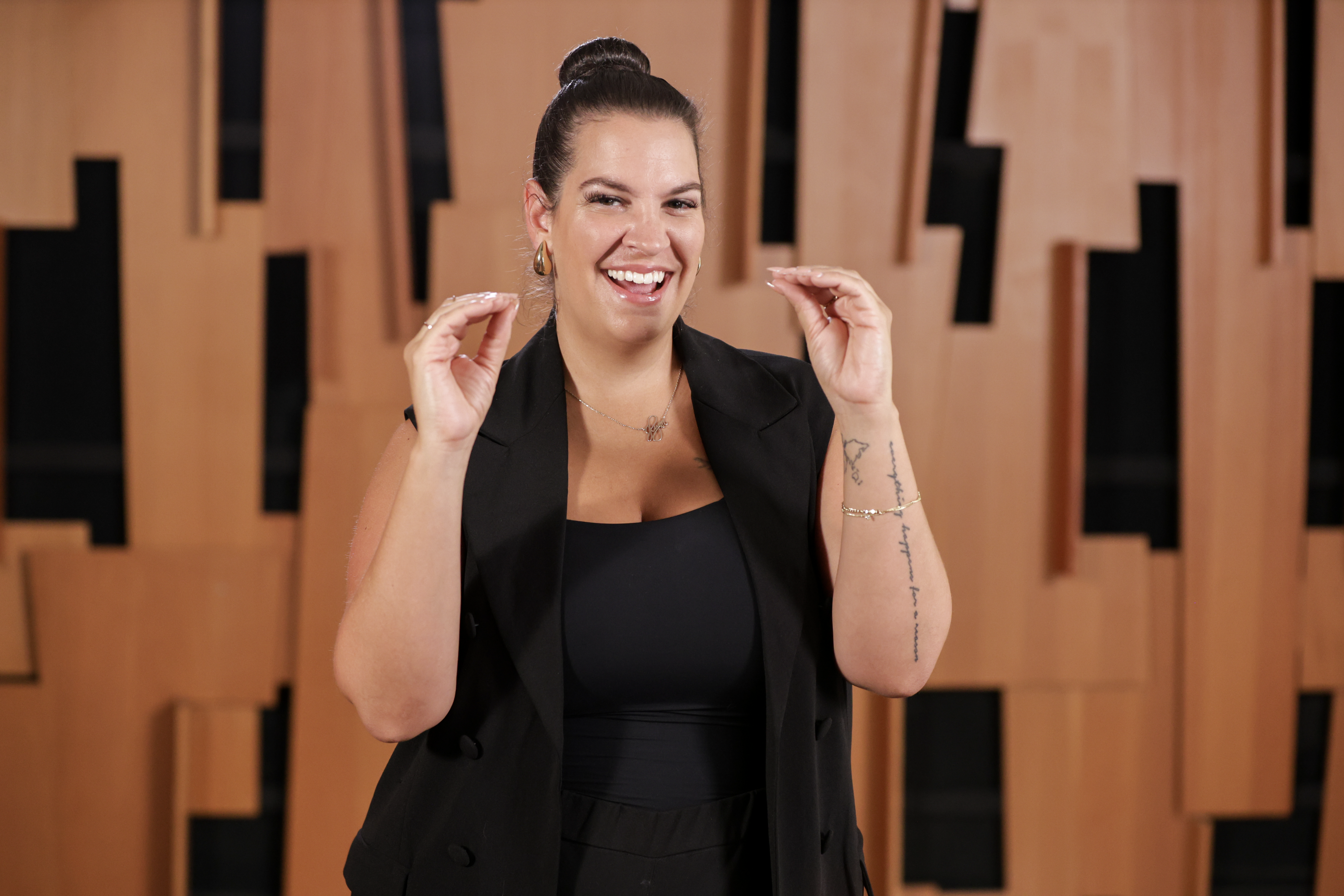 Jennifer Marfino in a black sleeveless pantsuit communicates in ASL in front of a wooden wall with black cut-outs.