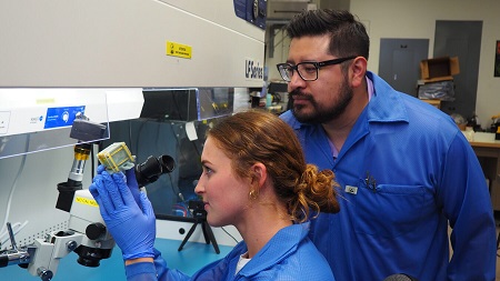 An image of two people in blue lab coats and gloves using equipment in a science lab