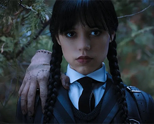 Jenna Ortega in a promo photo from the Netflix series “Wednesday” — dressed in a dark school uniform with braids and a severed hand on her shoulder