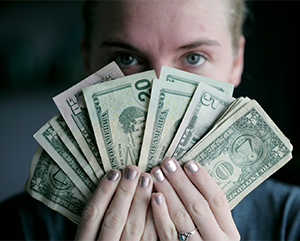 A close-up image of a blue-eyed blonde-haired woman holding a fan of U.S. dollars