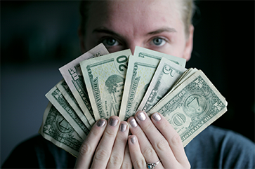 A close-up image of a blue-eyed blonde-haired woman holding a fan of U.S. dollars