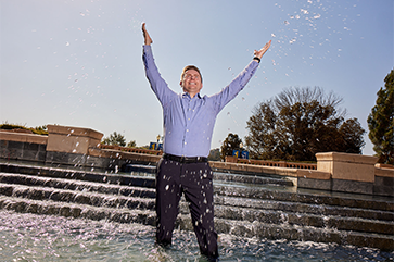 Andrew James-McClure with his hands pointing towards the sky as he dips his feet in UCLA's Shapiro Fountain.