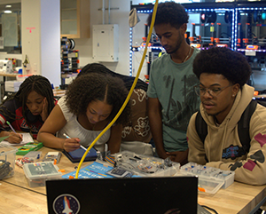 From left: Colene Agbo, Kayleen Speller, Yohannes Tefera and Ademole Turner write in notebooks at a lab table with pens and circuitry pieces.