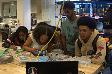 From left: Colene Agbo, Kayleen Speller, Yohannes Tefera and Ademole Turner write in notebooks at a lab table with pens and circuitry pieces.