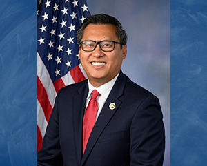 Representative Vince Fong, wearing a suit and a red tie, smiles for a portrait in front of an American flag.