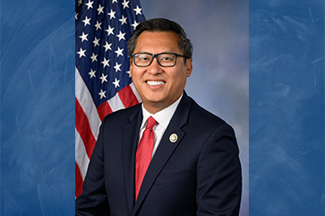 Representative Vince Fong, wearing a suit and a red tie, smiles for a portrait in front of an American flag.