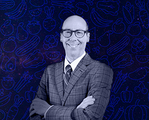 A black and white, head-and-shoulders headshot of Jack Bobo superimposed against a galaxy with blue outlines of constellation-shaped fruits and vegetables.