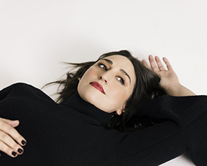 Sarah Bareilles in black lying on a white floor with left hand above her head