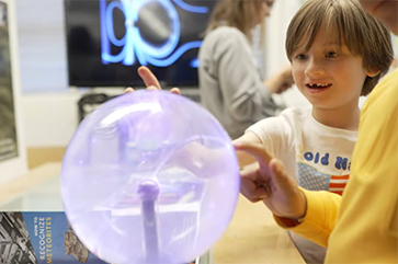 A young child touches a plasma globe, causing bright electric filaments. An adult with a yellow sleeve also engages with the globe, creating a shared moment of curiosity.