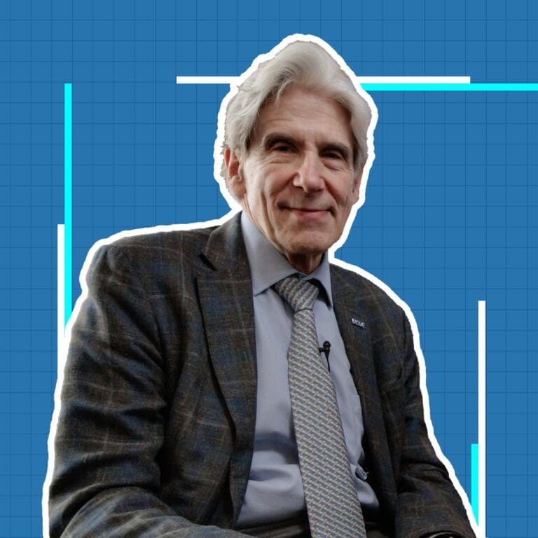 An older man with gray hair, wearing a plaid suit, dress shirt, and patterned tie, sits and smiles at the camera against a blue grid background with white and blue graphic lines.