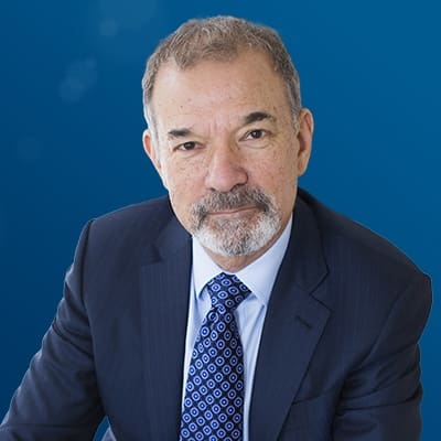 A middle-aged man with short gray hair, a trimmed beard, and mustache, wearing a navy suit, white shirt, and patterned blue tie, sits against a solid blue background.