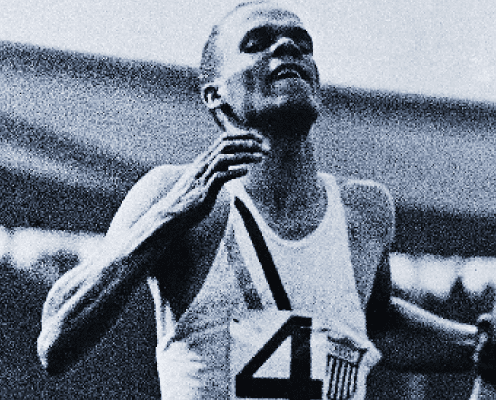 A male athlete wearing a tank top with the number 4 and a USA emblem crosses the finish line, arms raised and expression triumphant, during a race. The image is in black and white.