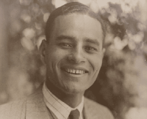 A young man in a suit and tie smiles warmly at the camera, with blurred foliage in the background. The image is sepia-toned, giving it a vintage feel.