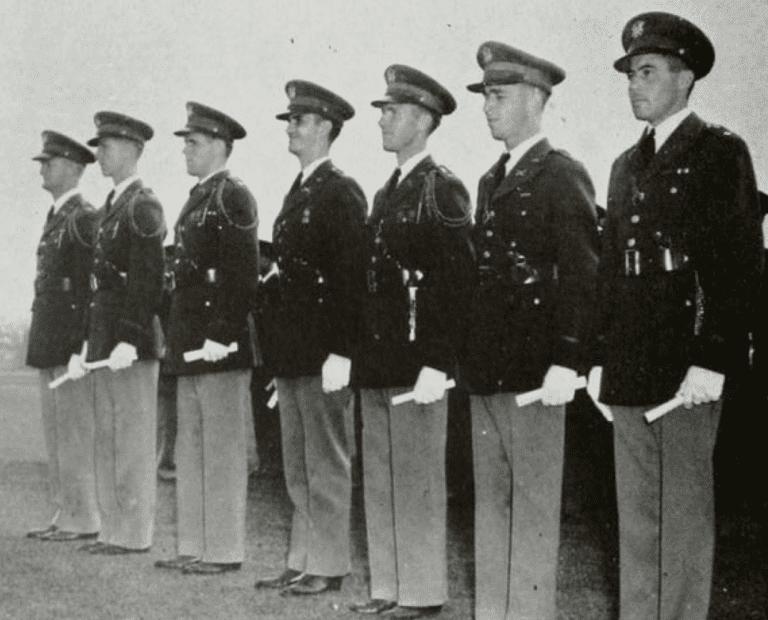 Seven men in military uniforms stand in a line outdoors, each holding a rolled document in their right hand. They appear to be part of a formal ceremony or graduation event.