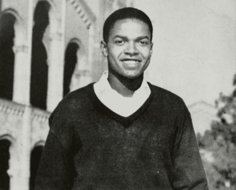 A young man wearing a dark sweater and white collared shirt smiles outdoors in front of a large, arched building. The photo is in black and white.
