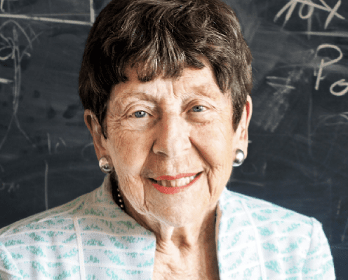 An older woman with short brown hair, pearl earrings, and a light-colored patterned jacket smiles in front of a chalkboard with writing and diagrams.