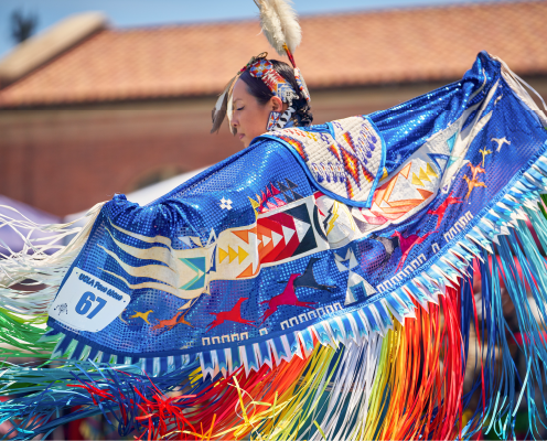 A dancer in vibrant traditional regalia with colorful fringe and patterns performs at a powwow. She wears a feathered headpiece and a blue shawl adorned with geometric and bird designs. A number “67” tag is visible on her outfit.