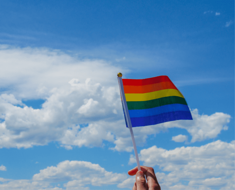 A hand holds up a small rainbow pride flag against a bright blue sky with scattered white clouds.