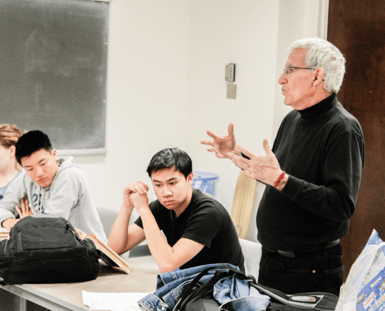 A man with gray hair and glasses speaks to a group of students seated at desks in a classroom. Two students listen attentively, while one looks down. A chalkboard is visible in the background.