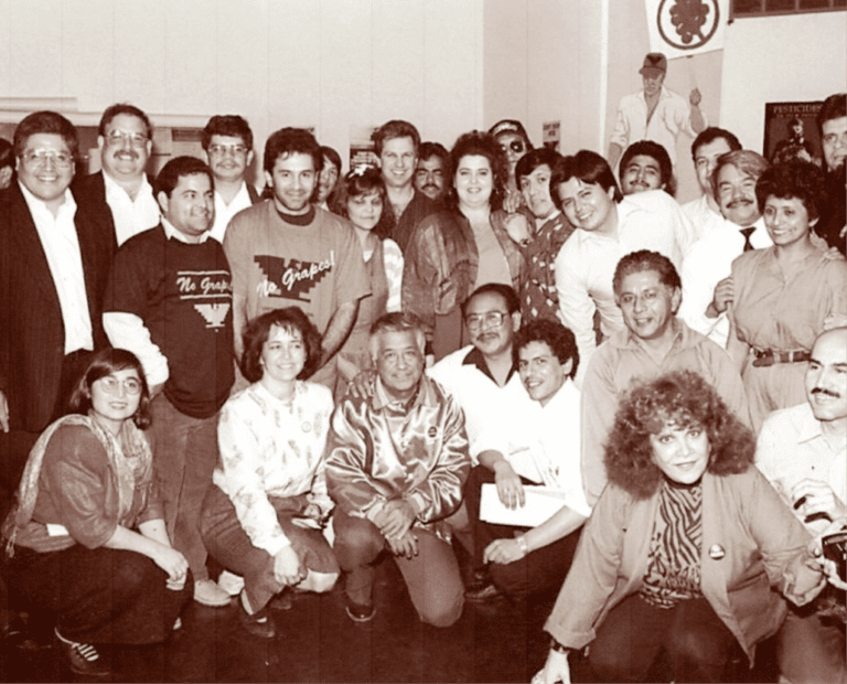 A large group of people, men and women, pose closely together indoors, smiling for the camera. The photo is in sepia tones and appears to be from the late 1980s or early 1990s.