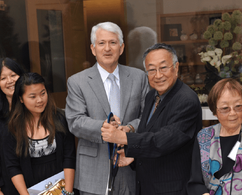 A group of people, including two men in suits holding large scissors, gather for a ribbon-cutting ceremony. Others stand nearby smiling, and potted plants are visible in the background.