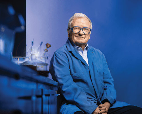 An elderly man with gray hair and glasses, wearing a blue lab coat, sits and smiles in a laboratory setting with scientific equipment visible on the counter beside him. The background is blue.