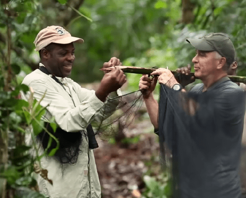 Two men stand in a lush, green forest, smiling while holding and untangling a fishing net together. Both wear outdoor clothing and hats, surrounded by dense vegetation.