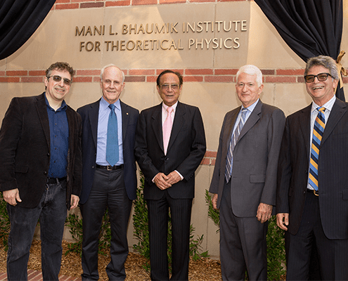 Five men in suits and jackets stand smiling in front of a building with a sign that reads Mani L. Bhaumik Institute for Theoretical Physics. Black curtains frame the sign above them.