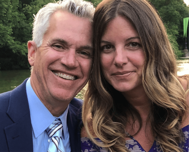 A man in a suit and tie and a woman in a purple dress smile closely together outdoors, with green trees and water in the background.