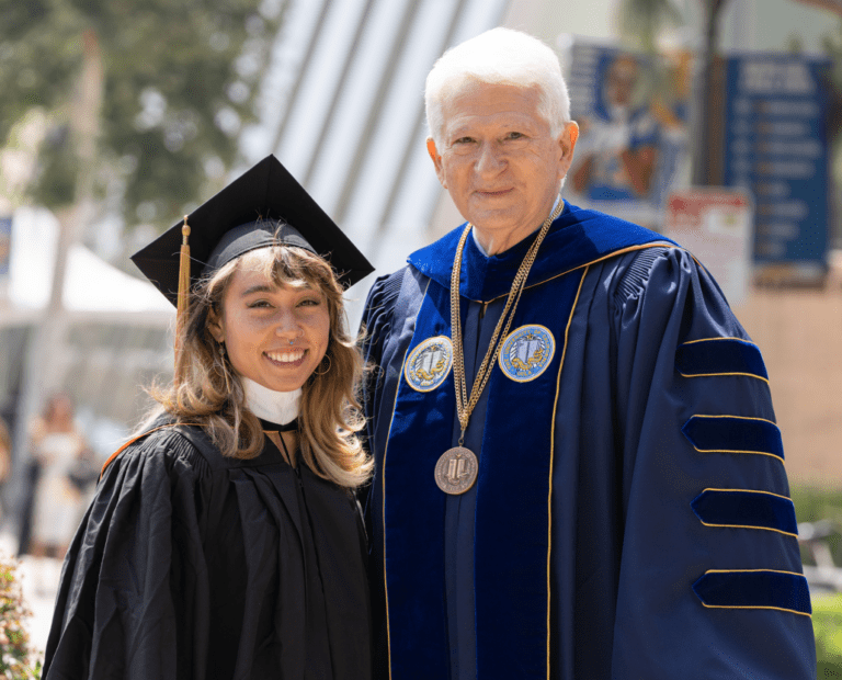 A young woman in a graduation cap and gown stands smiling next to an older man in academic regalia and a medallion, both posing outdoors on a sunny day.