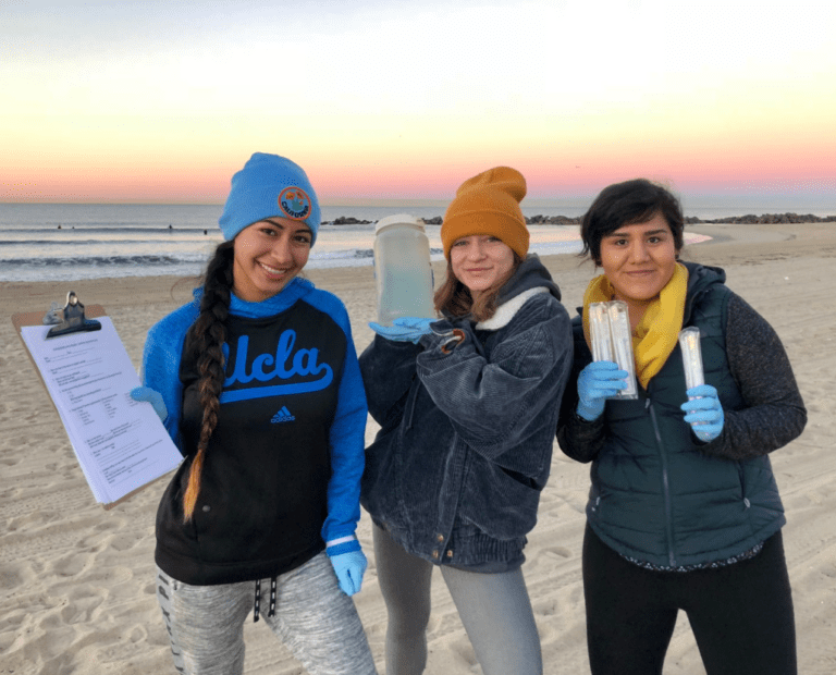Three young women stand on a sandy beach at sunset, smiling and holding scientific equipment: a clipboard, a jar of water, and test tubes. All wear gloves, warm hats, and casual winter clothing. The ocean is in the background.