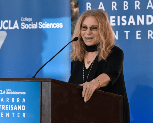 A woman with long blonde hair and glasses speaks at a podium with UCLA College Social Sciences and Barbra Streisand Center displayed on blue banners in the background.