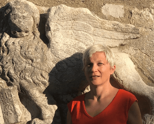 A person with short white hair wearing an orange top stands in sunlight in front of a weathered stone sculpture of a winged lion.