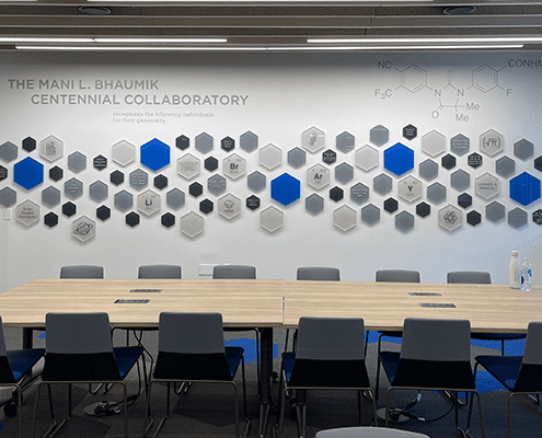 A modern meeting room with a wooden table, blue and gray chairs, and a wall decorated with hexagonal tiles featuring chemical symbols and names, labeled The Mani L. Bhaumik Centennial Collaboratory.