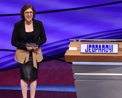 A woman in a black dress and tan blazer stands smiling on the Jeopardy! set, holding a Final Jeopardy! card beside the shows game podium with the Jeopardy! logo.